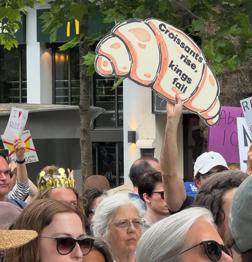 crowd and a sign shaped like a croissant that says croissants rise kings fall from the no kings rally in june