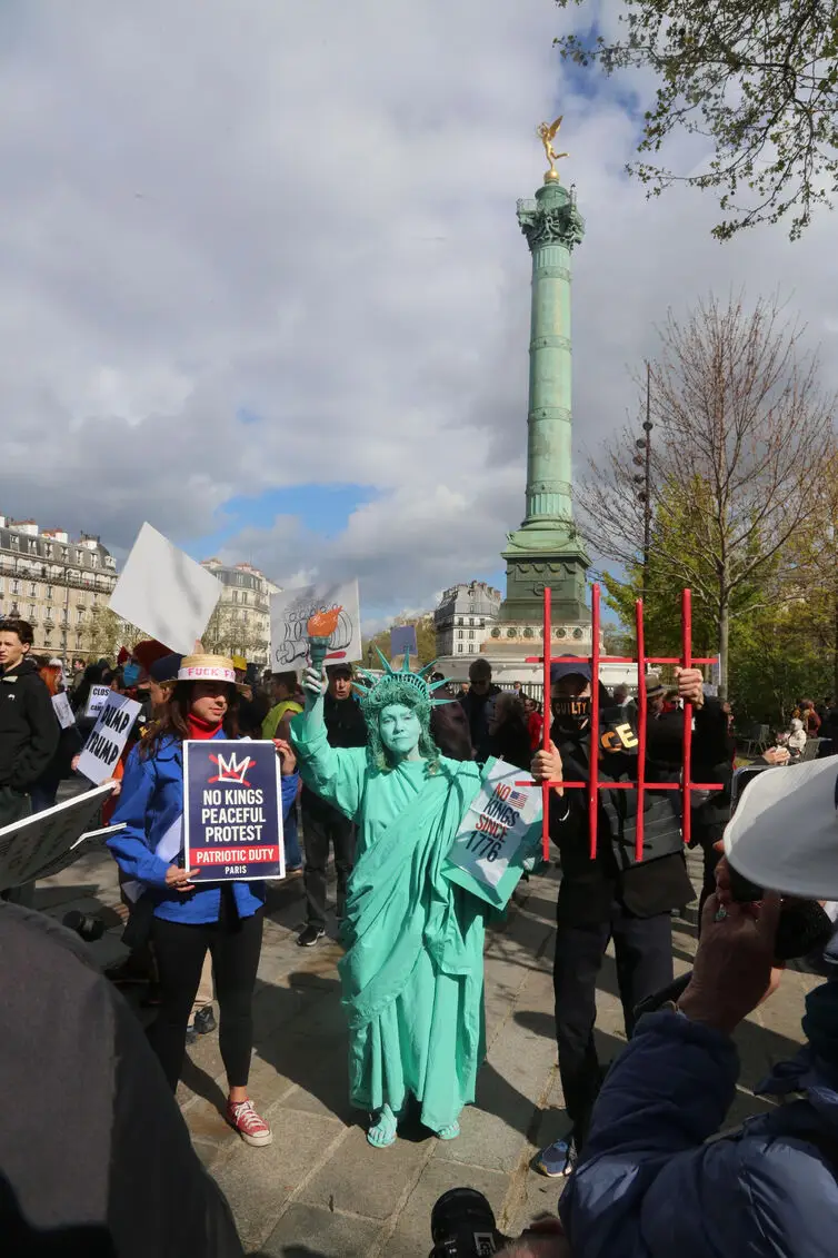 Photo show No Kings 3 rally at place de la Bastille in Paris, France. Photo show a man dressed as ICE behind bars, Lady Liberty, and a woman holding a No Kings 3 sign. Photo credit: Owen Franken