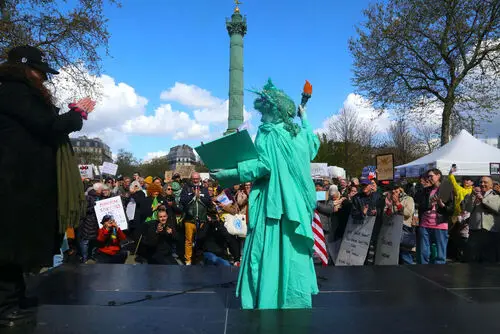 Image shows the Paris No Kings 3 rally on march 28th 2026. In the foreground a woman dressed as Lady Liberty is seen from behind. IN the mid ground is the crowd and in the background is the Ma Bastille Tower. Photo credit is Owen Franken